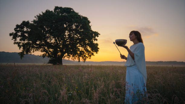 A mature Caucasian woman engages in a tranquil meditation, playing a Tibetan singing bowl in a meadow, as the day's last light paints the scene serene.